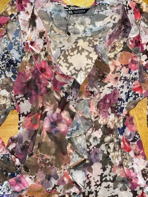 Blommig volangblus från Zara - Säljer en transparent blus från Zara med blommigt mönster i rosa, lila, brun och blå toner. Blusen har långa ärmar, v-ringning och snygga volangdetaljer framtill och längs axlarna. Perfekt för dig som gillar färg och romantiska vibes.