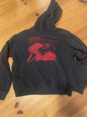 Svart Deftones hoodie från H&M Divided XS - Svart hoodie från H&M Divided i storlek XS med stor röd Deftones-tryck på framsidan. Tröjan har huva och långa ärmar, tillverkad i mjuk bomull. Snyggt grafiskt motiv och cool streetstil för dig som gillar bandmerch.