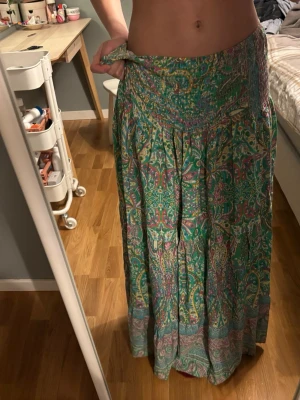 Marta du chateau kjol - Säljer en flowy långkjol med grönt som basfärg och färgglatt paisleyinspirerat mönster i lila, gult och rosa. Kjolen har bred smock i midjan och är gjord i ett tunt, mjukt tyg som känns luftigt och skönt. Perfekt för dig som gillar boho-vibbar och färg! Tyvärr för stor för mig, passar nog s/m och uppåt!