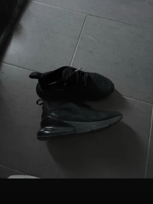 Svarta Nike Air Max 270 sneakers - Säljer ett par helsvarta Nike Air Max 270 sneakers med snygg, sportig design. Skorna har mesh-ovandel för bra ventilation, synlig Air-enhet i hälen och snörning för perfekt passform. Perfekta för dig som gillar streetwear och vill ha bekväma skor till vardagen.