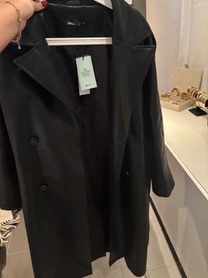 Svart lång skinnkappa från Gina Tricot - Säljer en svart lång skinnkappa från Gina Tricot med klassisk krage och knappar framtill. Kappan har en rak och oversized passform, perfekt för dig som vill ha en stilren look. Materialet är syntetiskt skinn som ger en cool vibe.