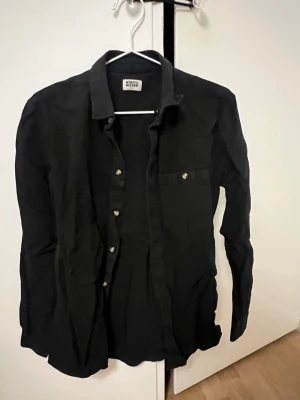 Svart skjorta från Weekday i bomull - Klassisk svart skjorta från Weekday med lång ärm och knappstängning framtill. Skjortan har en bröstficka med knapp och är tillverkad i mjuk bomull. Perfekt att styla lager på lager eller bära som den är.