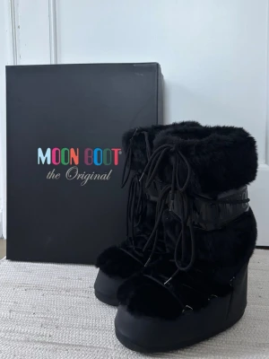 Svarta Moon Boots med fuskpäls - Säljer ett par svarta Moon Boots med fluffig fuskpäls och snörning framtill. Skorna har hög platåsula och bred passform, perfekta för kalla vinterdagar. Klassisk Moon Boot-logga i blank plast runt skaftet och riktigt mysig vibe.