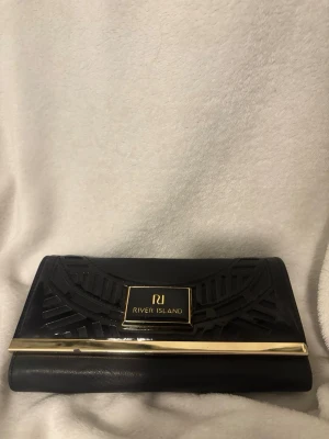 Svart plånbok från River Island - Snygg svart plånbok från River Island med guldfärgade detaljer och coolt geometriskt mönster på framsidan. Plånboken har flera fack för kort, mynt och sedlar samt en genomskinlig ficka för ID. Stängs med tryckknapp och metallspänne.