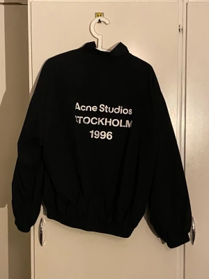 Svart vindjacka från Acne Studios - Säljer en svart vindjacka från Acne Studios med tryck 'Acne Studios STOCKHOLM 1996' på ryggen. Jackan har hög krage, dragkedja framtill och elastiska muddar vid ärmar och midja. Enkel och clean design som passar till det mesta.