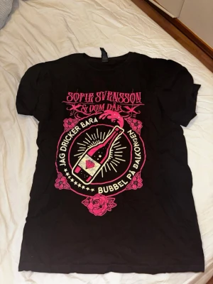 Svart t-shirt Sofie Svensson tryck - Svart t-shirt med färgstarkt rosa och vitt tryck framtill. Motiv med texten 'Jag dricker bara bubbel på balkongen' och en bubbel-flaska omgiven av rosor. Klassisk rund hals och korta ärmar. Perfekt för dig som gillar statement-plagg och coola bandmotiv.