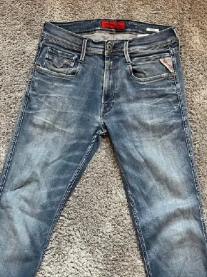 Replay Anbass blå jeans, storlek 31 - Säljer ett par Replay Anbass jeans i klassisk blå tvätt med snygga slitningar och detaljerade sömmar. Modellen har normal passform, fem fickor och dragkedja. Perfekta för dig som gillar en avslappnad men ändå stilren look.