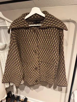 Brun och beige rutig jacka - Unik jacka i brun och beige med rutigt mönster och stora knappar framtill. Jackan har bred krage och är långärmad, tillverkad i ett stickat material som ger en mysig och cool vibe. Perfekt för dig som gillar retro och vill sticka ut.