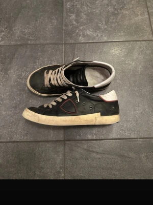 Philippe Model svarta sneakers i skinn - Svarta sneakers från Philippe Model med vita detaljer och vit sula. Skorna har snörning, rund tå och en ikonisk sköld på sidan med röd kant. Ovandelen är i skinn och hälen är vit med diskret logga. Klassisk och stilren modell som passar till mycket.