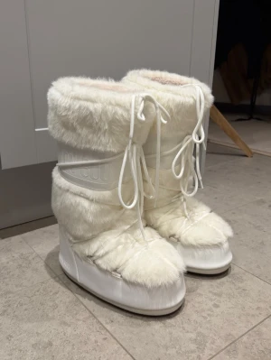 Vita Moon Boots med fuskpäls - Säljer ett par vita Moon Boots med fluffig fuskpäls och snörning framtill.  Nyskick! Inga fläckar. Nypris: 3315kr