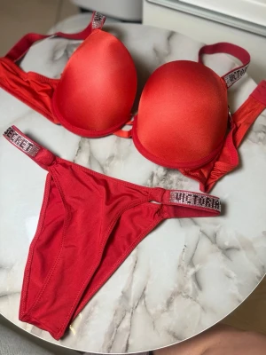 Röd bh och trosa från Victoria's Secret - Snyggt rött set från Victoria's Secret med vadderad bh och matchande trosa. Axelbanden och sidorna på trosan har glittriga detaljer med logga i strass. Materialet är mjukt och stretchigt, perfekt för dig som vill ha något extra. Setet har en modern och ungdomlig vibe.