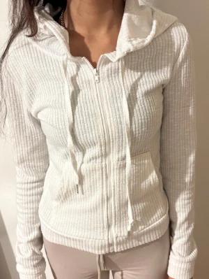 Vit ribbad hoodie med dragkedja - Säljer en vit ribbad hoodie med dragkedja framtill och huva. Tröjan har långa ärmar, snörning i huvan och två fickor framtill. Perfekt för en chill och avslappnad stil.
