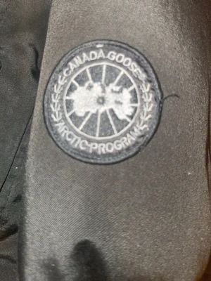Svart dunjacka från Canada Goose - Säljer en svart dunjacka från Canada Goose med klassisk patch på ärmen och diskreta detaljer. Jackan har dragkedja och knappar framtill samt ribbade muddar vid ärmsluten. Perfekt för kalla dagar och riktigt snygg till alla outfits.