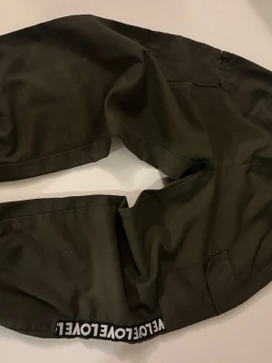 Mörkgröna cargopants med LOVE-detalj - Säljer ett par mörkgröna cargopants i bomull med cool svartvit LOVE-text längs benslutet. Byxorna har fickor på sidorna och avslappnad passform. Perfekta för dig som gillar streetwear och vill ha något bekvämt men ändå snyggt.