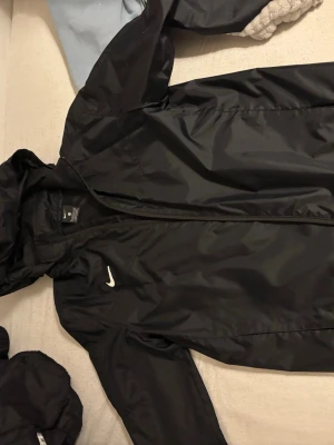Svart Nike vindjacka med huva - Säljer en svart vindjacka från Nike med huva och dragkedja framtill. Jackan har en enkel och clean look med Nike-logga på bröstet. Tillverkad i lätt polyester, perfekt för blåsiga dagar. Långärmad modell med elastiska muddar.