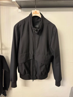 Svart bomberjacka från J.Lindeberg - Säljer en svart bomberjacka från J.Lindeberg i sportigt, lätt material från Loro Piana Storm System. Jackan har hög krage, ribbade muddar och dragkedja framtill. Snygg minimalistisk look med diskreta fickor och clean passform.