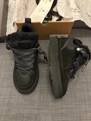  UGG lowmel - Säljer ett par svarta boots från UGG med grov sula och fluffig insida. Skorna har breda, mönstrade snören i svart och grått, samt detaljer i mocka och mesh. Perfekta för kalla dagar med sin varma insida och robusta look.