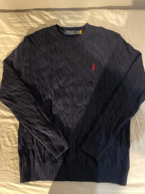 Mörkblå kabelstickad tröja Polo Ralph Lauren - Kabelstickad mörkblå tröja från Polo Ralph Lauren med klassisk röd logga broderad på bröstet. Tröjan har rund halsringning, ribbade muddar och är långärmad. Perfekt för dig som gillar stilrena och tidlösa plagg.