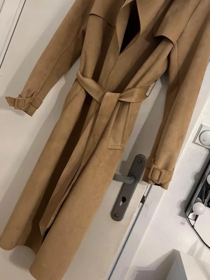 Beige trenchcoat med bälte ZARA - Snygg beige trenchcoat med bälte i midjan och spännen vid ärmsluten. Jackan har en klassisk lång siluett och är tillverkad i mockaliknande material som ger en lyxig känsla. Perfekt för dig som vill ha en stilren och trendig look.