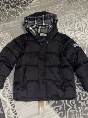 Svart pufferjacka från Burberry - Säljer en svart pufferjacka från Burberry med klassiskt rutigt foder i huvan. Jackan har dragkedja och knappar framtill, två fickor med dragkedja och en stor Burberry-patch på ärmen. Perfekt för kalla dagar och riktigt snygg streetstil.