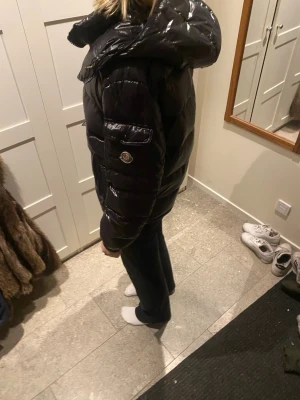 Svart pufferjacka från Moncler - Säljer en svart pufferjacka från Moncler med glansig finish och huva. Jackan har Moncler-logga på ärmen, dragkedja framtill och coolt serietidningsmotiv på insidan. Perfekt för dig som vill ha en varm och snygg jacka med exklusiv känsla.