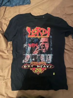 Lordi Get Heavy 20-års t-shirt - Svart t-shirt med stort Lordi-tryck fram och texten 'Get Heavy samt bandmedlemmarnas ansikten. Baksidan har texten 'I Got Heavy Again' och en mask. Perfekt för dig som älskar metal och vill sticka ut med en fet bandtröja.