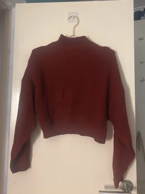 Vinröd croppad stickad tröja - Säljer en vinröd croppad stickad tröja med hög hals och långa ärmar. Tröjan har en ribbad struktur och är perfekt för dig som gillar en trendig och mysig stil. Passar till både jeans och kjol för en snygg look. Storlek L men passar S/M. Aldrig använd