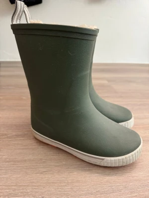 Gröna gummistövlar från Tretorn - Mörkgröna gummistövlar från Tretorn med vitt foder och breda randiga dragflikar upptill. Stövlarna har en vit sula och reflekterande detalj baktill. Perfekta för regniga dagar och håller fötterna torra.