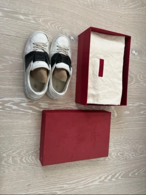 Valentino Garavani vita sneakers - Säljer ett par vita Valentino Garavani sneakers med svart bred läderdetalj över vristen och ikoniska nitar bak på hälen. Skorna har snörning, rund tå och platt sula. Kommer med original röd box och dustbag. Perfekta för dig som vill ha en lyxig och edgy look.