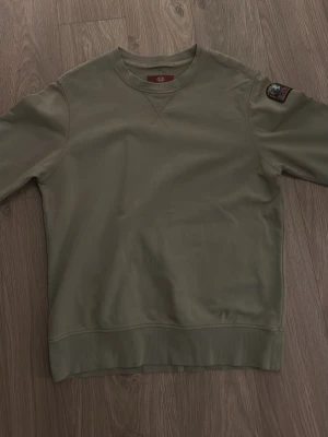Olivgrön sweatshirt från Parajumpers - Säljer en olivgrön sweatshirt från Parajumpers med klassisk rund hals och ribbade muddar. Tröjan har en patch med logga på ena ärmen och är perfekt för dig som gillar enkel men snygg stil.