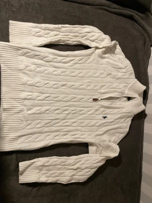 Vit stickad polotröja Polo Ralph Lauren - Säljer en vit stickad polotröja från Polo Ralph Lauren med klassiskt kabelstickat mönster och half zip-dragkedja med läderdetalj. Tröjan har ribbade muddar, hög krage och den ikoniska loggan broderad på bröstet. Perfekt för höst och vinter! Passar även mig som har storlek S