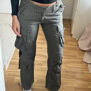 Gröna cargopants med fickor - Säljer ett par gröna cargopants med flera stora fickor på sidorna och detaljer som knappar och snörning. 