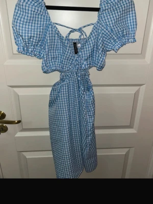 Blåvit rutig omlottklänning Divided - Supersöt blåvit rutig klänning från Divided med puffärmar, omlottdetalj och snörning fram. Klänningen har cut-out i midjan och resår i ärmslut och midja för en skön passform. Perfekt för sommarens alla tillfällen!