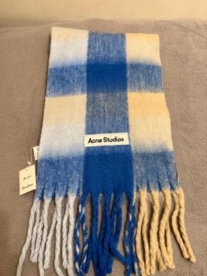 Halsduk Acne Studios - Snygg halsduk från Acne Studios med stort rutmönster i blått, beige och vitt. Mjuk och fluffig känsla med långa tvinnade fransar i matchande färger. Märkeslapp framtill. Perfekt accessoar för att lyfta din look.