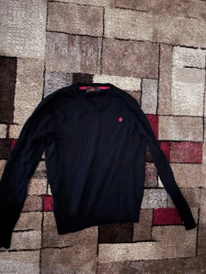 Svart merinoullströja Morris S - Klassisk svart tröja från Morris i 100% merinoull. V-ringad modell med lång ärm och diskret röd logga broderad på bröstet. Perfekt till lager på lager och funkar året runt. Snygg röd detalj på insidan av kragen.