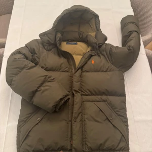 Olivgrön pufferjacka Polo Ralph Lauren - Säljer en olivgrön pufferjacka från Polo Ralph Lauren med huva och orange broderad logga på bröstet. Jackan har dragkedja, tryckknappar och två stora fickor framtill. Fodret är beige och jackan är riktigt fluffig och varm, perfekt för kalla dagar. Pris kan även diskuteras.