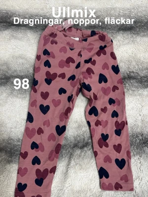 Rosa ullmix leggings med hjärtan - Söta rosa leggings i ullmix med tryckta hjärtan. Mjuka och stretchiga med resår i midjan, perfekta för lek och chill. Hjärtprinten ger en lekfull vibe och materialet är skönt mot huden.🌸Ullmix. Dragningar. Noppor, fläckar. Storlek 98