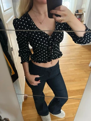 Svart prickig croppad blus - Säljer en svart croppad blus med vita prickar och volangdetaljer. Toppen har v-ringning, knappar framtill och långa ärmar med lätt puff. Perfekt för dig som gillar en lekfull och trendig stil.