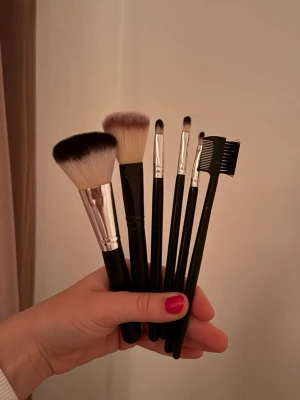 Set med sminkborstar och brynkam - Säljer ett set med sex sminkborstar och en ögonbrynskam. Borstarna har svarta handtag och syntetiska borst i vitt och brunt. Perfekt för applicering av puder, ögonskugga och detaljer. Brynkammen har både borste och kam för att fixa brynen. 