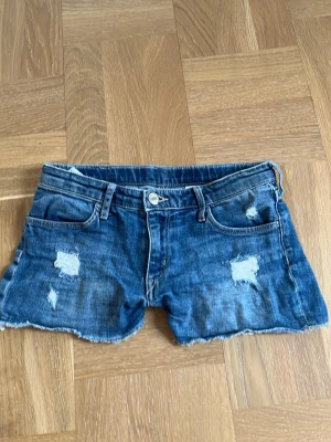 Blå denimshorts från Denim - Säljer ett par blå low waisted jeansshorts från Denim med råa kanter och slitna detaljer framtill. Klassisk femficksmodell med knapp och dragkedja. Perfekta till sommaren och ger en avslappnad vibe.