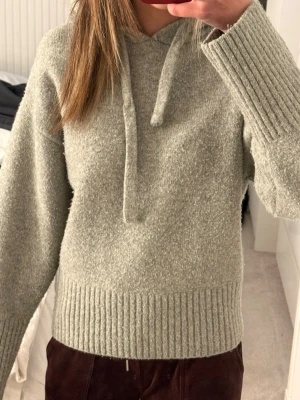 Ljusgrå stickad hoodie med huva - Mysig ljusgrå stickad hoodie med huva och snörning. Tröjan har breda ribbade muddar vid ärmslut och nederkant samt lång ärm. Perfekt för kyliga dagar och enkel att matcha till olika outfits.