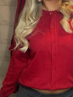 Röd kofta från Jacett, storlek 38/40 - Klassisk röd kofta från Jacett i storlek 38/40. Koftan har rund halsringning, knappar hela vägen fram och två fickor framtill. Tillverkad i mjukt stickat material och har långa ärmar. Perfekt att slänga över en topp för en chill look.