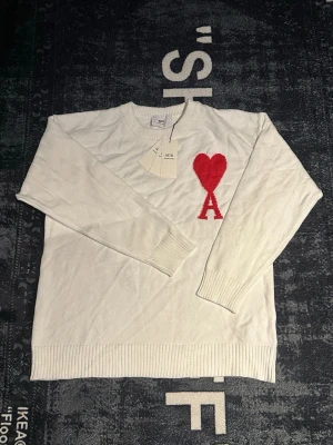Vit stickad tröja från Ami med hjärta - Säljer en vit stickad tröja från Ami Alexandre Mattiussi med röd hjärtdetalj och bokstaven A på bröstet. Tröjan har rund halsringning, ribbade muddar och är långärmad. Perfekt för dig som gillar stilrena plagg med en twist.