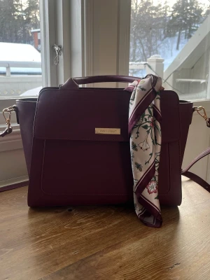 Vinröd axelväska från Anna Field - Säljer en vinröd axelväska från Anna Field i stilren design. Väskan har guldfärgade detaljer, ett handtag upptill och avtagbar axelrem. Utsidan är i syntetmaterial och insidan har flera fack. En snygg scarf med blommigt mönster i vitt, grönt och rött följer med. Väskan är aldrig använd. Endast upp plockad ur förpackning 