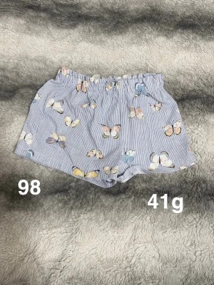 Randiga shorts med fjärilar - Söta shorts med smala blå och vita ränder och färgglada fjärilar tryckta över hela tyget. De har resår i midjan för skön passform och är perfekta för varma dagar. Lätta och luftiga, passar perfekt till sommaren.🌸 Fint använt skick. Storlek 98 