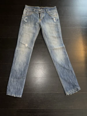 Blå slitna jeans från Nudie Jeans - Säljer ett par blå jeans från Nudie Jeans med slitningar och ljusa tvättade partier. Klassisk femficksmodell med raka ben och kontrastsömmar på bakfickorna. Jeansen har normal passform och är tillverkade i bomull.