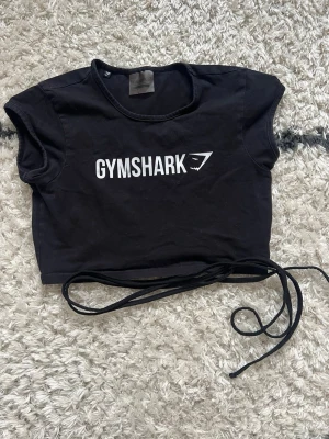 Gymshark Topp  - Svart träningstopp från Gymshark med vit logga framtill. Toppen har korta ärmar och långa knytband som kan lindas runt midjan för en snygg detalj 🖤