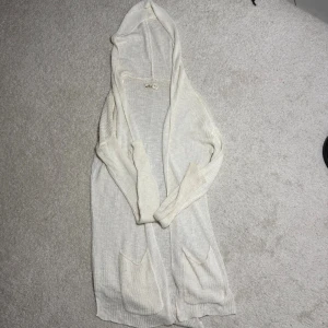 Vit lång kofta med huva från Hollister - Lång, öppen kofta från Hollister i vit stickad bomullsmix. Tröjan har huva, två stora fickor framtill och långa ärmar. Perfekt för lager på lager och chill dagar. Enkel och clean design som passar till det mesta.