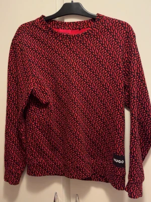 Röd och svart långärmad tröja HUGO - Säljer en snygg långärmad tröja från HUGO med rött och svart abstrakt mönster över hela plagget. Tröjan har rund halsringning och en liten HUGO-logga nertill. Perfekt för dig som vill sticka ut med en cool design.