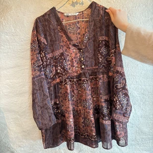 Mönstrad kortklänning från Zara - Superfin kortklänning från Zara med paisleyinspirerat mönster i lila, brun och rosa toner. Klänningen har v-ringning med knappar framtill, långa ärmar och ett luftigt, flowy material. Perfekt för dig som gillar boho-vibe och snygga detaljer.